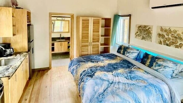 A bedroom with a wooden ceiling, a bed with a blue and white patterned bedspread, a wooden wardrobe, and a kitchenette area. - Conch Casitas - King or 2 Singles, AC, Shared Pool (Roatán)