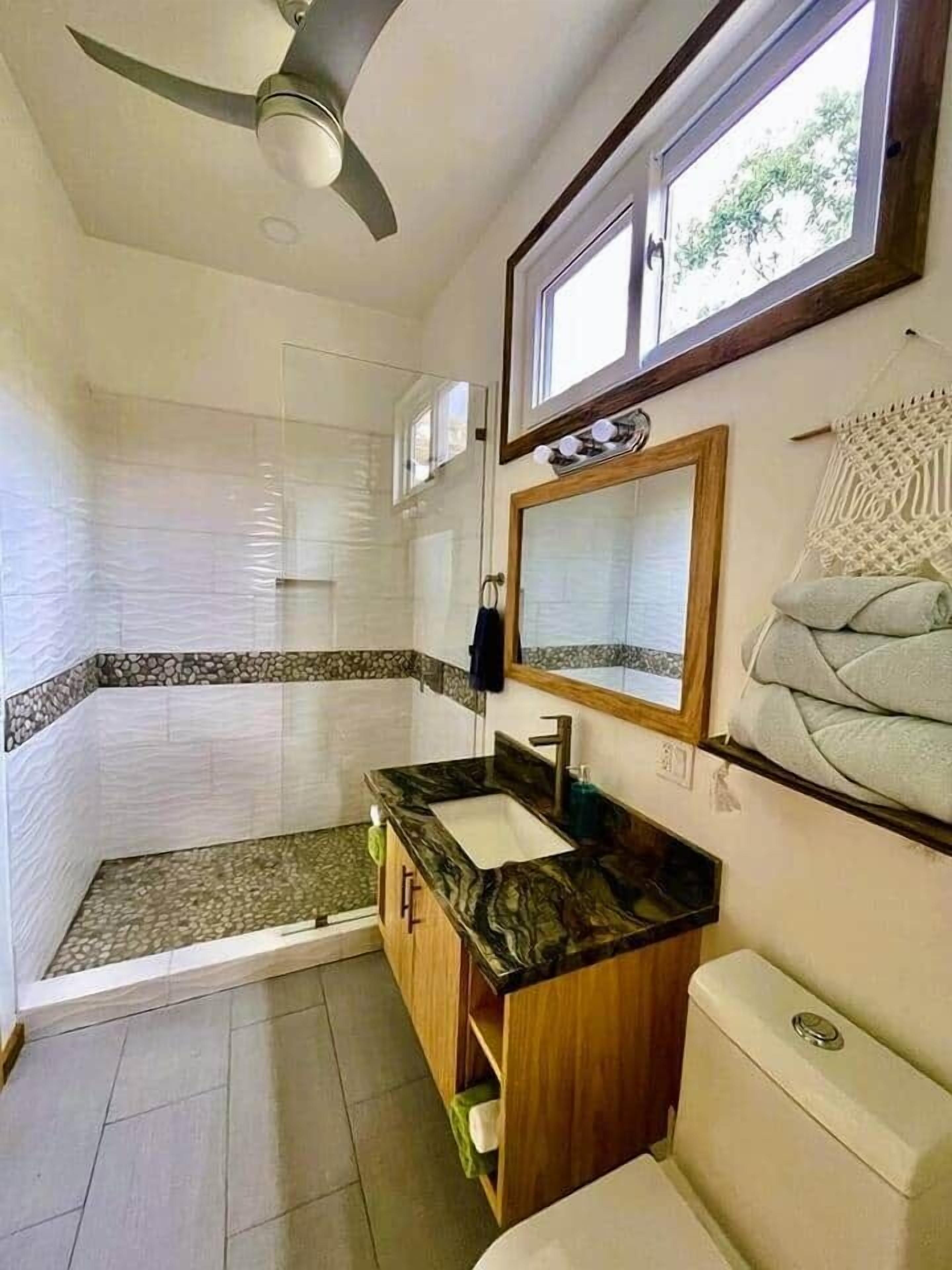 A bathroom with a shower, a vanity with a sink, a toilet, a ceiling fan, and a window.