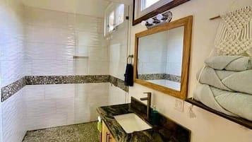 A bathroom with a shower, a vanity with a sink, a toilet, a ceiling fan, and a window.