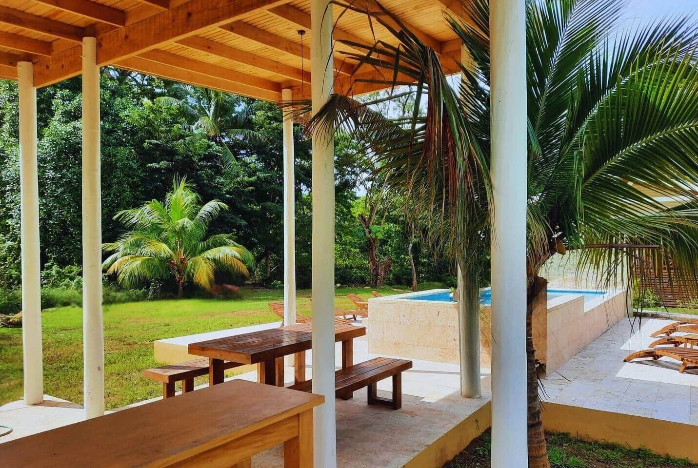 An outdoor patio with wooden tables and benches, surrounded by lush greenery and a swimming pool.