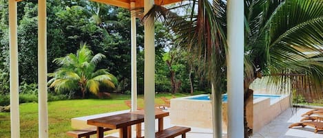An outdoor patio with wooden tables and benches, surrounded by lush greenery and a swimming pool.