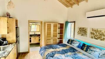 A bedroom with a wooden ceiling, a bed with a blue and white patterned bedspread, a wooden wardrobe, and a kitchenette area.