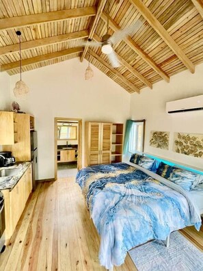 A bedroom with a wooden ceiling, a bed with a blue and white patterned bedspread, a wooden wardrobe, and a kitchenette area. - Conch Casitas - King Bed, AC, Shared Pool (Roatán)