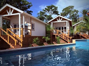 A poolside view of a resort with two wooden cabins, each with a staircase leading to a deck. - Conch Casitas - King Bed, AC, Shared Pool (Roatán)