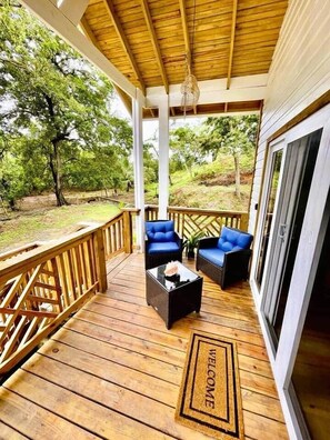 A wooden deck with two blue chairs and a coffee table, a welcome mat, and a view of trees. - Conch Casitas - King Bed, AC, Shared Pool (Roatán)
