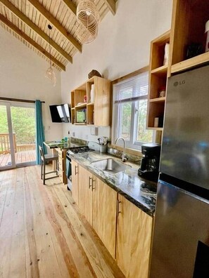 A kitchen with wooden cabinets, a stainless steel refrigerator, and a window overlooking greenery. - Conch Casitas - King Bed, AC, Shared Pool (Roatán)