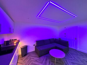 A modern living space with a sectional sofa, a small round table, and a kitchen area with cabinets. - 'the Garden Room' Studio, Sleeps 2 - Wifi, Parking (Edinburgh)