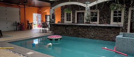 Una piscina interior con pared de piedra, techo con tragaluces y zona de estar con sillas y mesas.