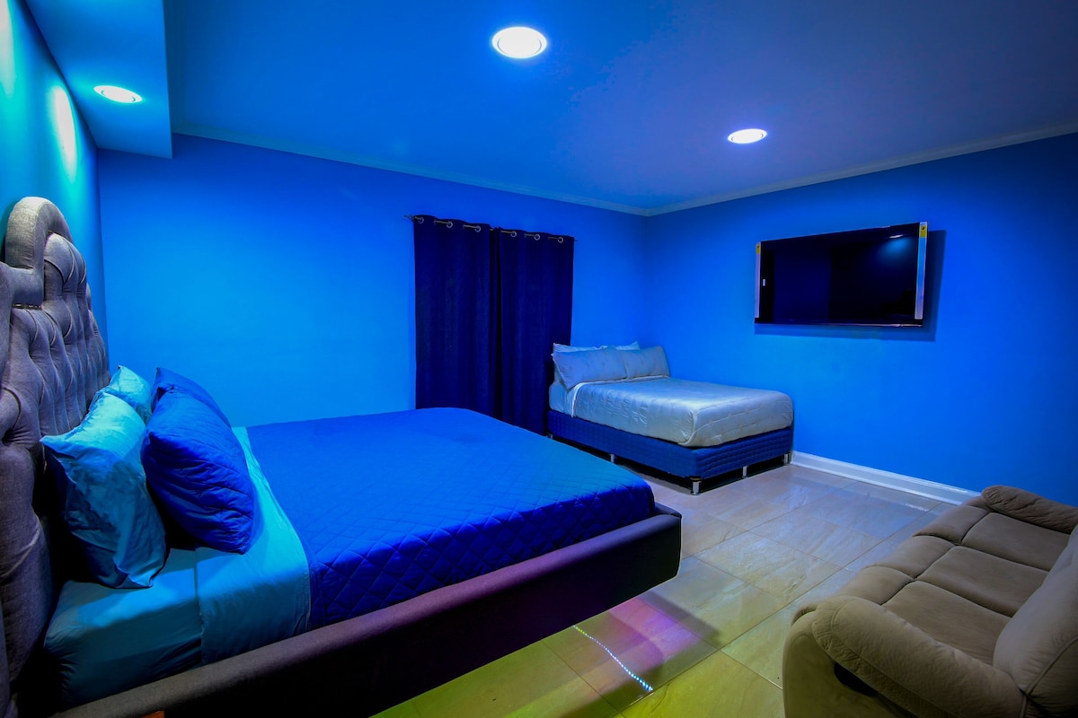Un dormitorio con una cama grande, una cama más pequeña, un televisor montado en la pared y una silla.