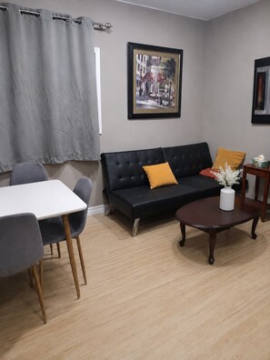 Ein Wohnzimmer mit einer schwarzen Couch, einem runden Couchtisch und einem Essbereich mit einem weißen Tisch und grauen Stühlen.
