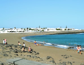 Ein Strand mit badenden Menschen, weißen Gebäuden im Hintergrund und einem klaren Himmel.