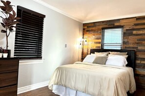 Un dormitorio con cama, mesita de noche, planta en maceta, ventana con persianas y pared con diseño de paneles de madera.