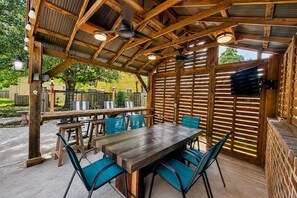 Un patio al aire libre con una mesa y sillas de madera, un televisor montado en la pared y un área cubierta con techo metálico.
