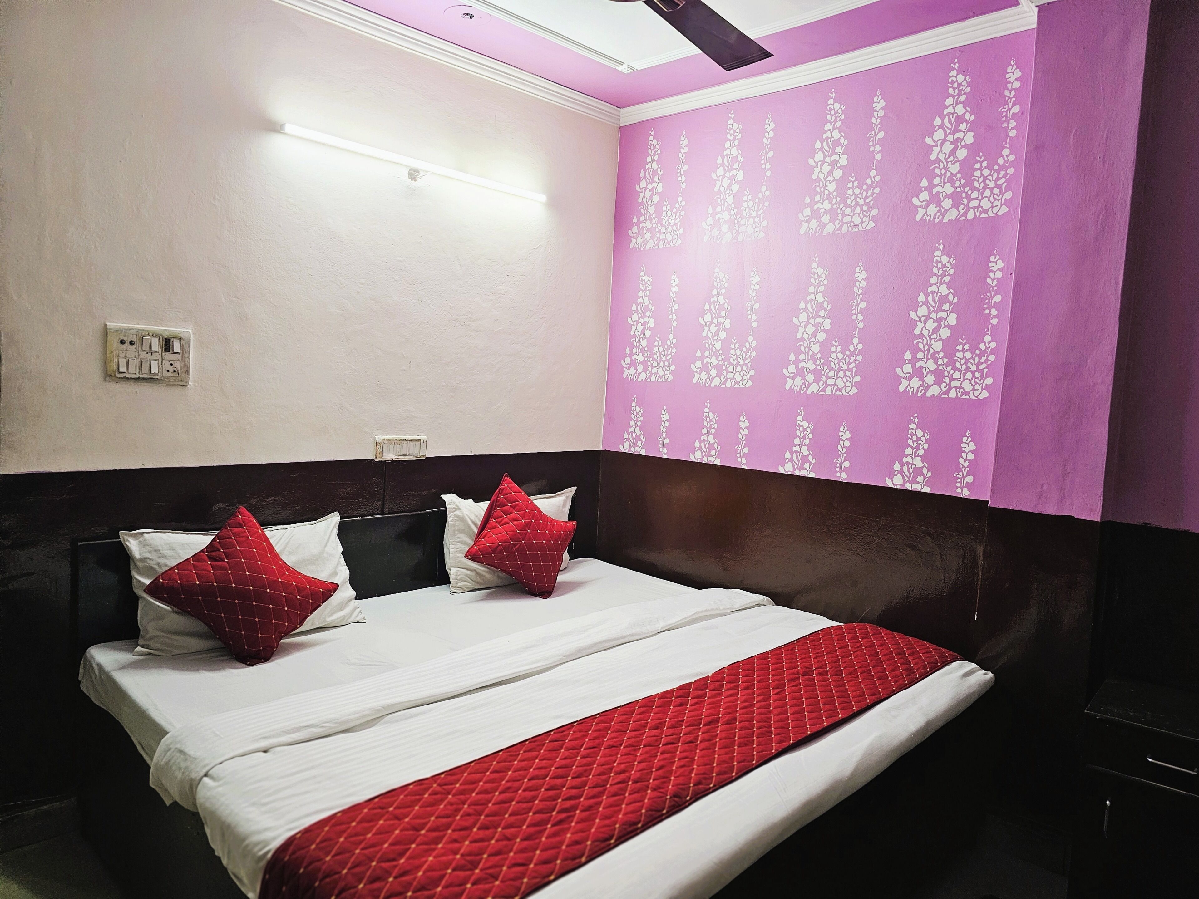 Une chambre d’hôtel avec un lit, une tête de lit, un mur recouvert d’un papier peint à motifs floraux et un luminaire au plafond.