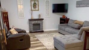 A living room with a fireplace, a flat-screen TV, a grey sectional sofa, and a black recliner. - Serene Escape. Tropical Gem. 
Cozy Chic Condo, 
Shared space Pvt. Bed/Bath w AC (Eustis)