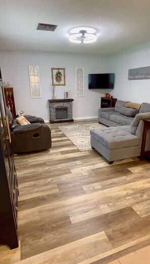 A living room with a fireplace, a flat-screen TV, a grey sectional sofa, and a black recliner. - Serene Escape. Tropical Gem. 
Cozy Chic Condo, 
Shared space Pvt. Bed/Bath w AC (Eustis)