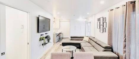 A modern living room with a sectional sofa, a flat-screen TV mounted on the wall, and a dining area with a wooden table and chairs.