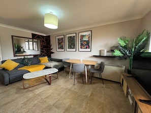 A modern living room with a sofa, a coffee table, and a dining table with chairs. There are framed pictures on the wall, a large plant, and a radiator. - Brixton Apartment with Stylish Touches (London)