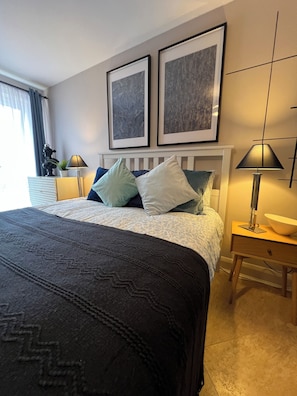 A hotel room with a bed, two framed artworks on the wall, a nightstand with a lamp, and a window with curtains. - Brixton Apartment with Stylish Touches (London)