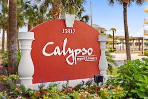 A red sign with 'Calypso' and 'resort & towers' text, surrounded by greenery and palm trees. - Enjoy Luxe King Mattress after Iron Man 💪Calypso II beachfront 19th Floor View! (Panama City Beach)