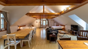 A spacious attic room with a high ceiling, wooden beams, a dining table, two beds, a sofa, and a television. - Auberge aux petits oiseaux (BAIE SAINT PAUL)