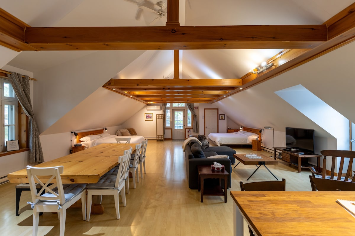 A spacious attic room with a high ceiling, wooden beams, a dining table, two beds, a sofa, and a television.