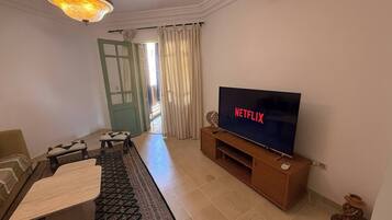 Een woonkamer met een televisie, een bank, een salontafel, een kamerbreed tapijt, een kroonluchter en een deur met gordijnen.