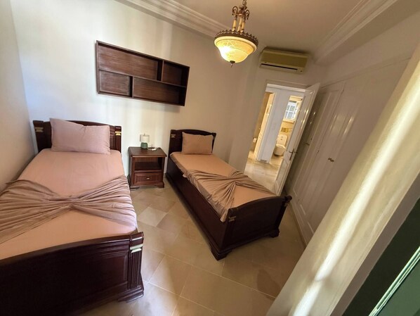 A hotel room with two beds, a nightstand, a lamp, a ceiling light, and a door leading to a bathroom.