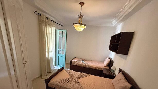A bedroom with two beds, a chandelier, a window with curtains, and a wall-mounted cabinet.