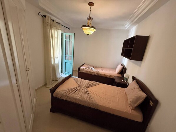 A bedroom with two beds, a chandelier, a window with curtains, and a wall-mounted cabinet.