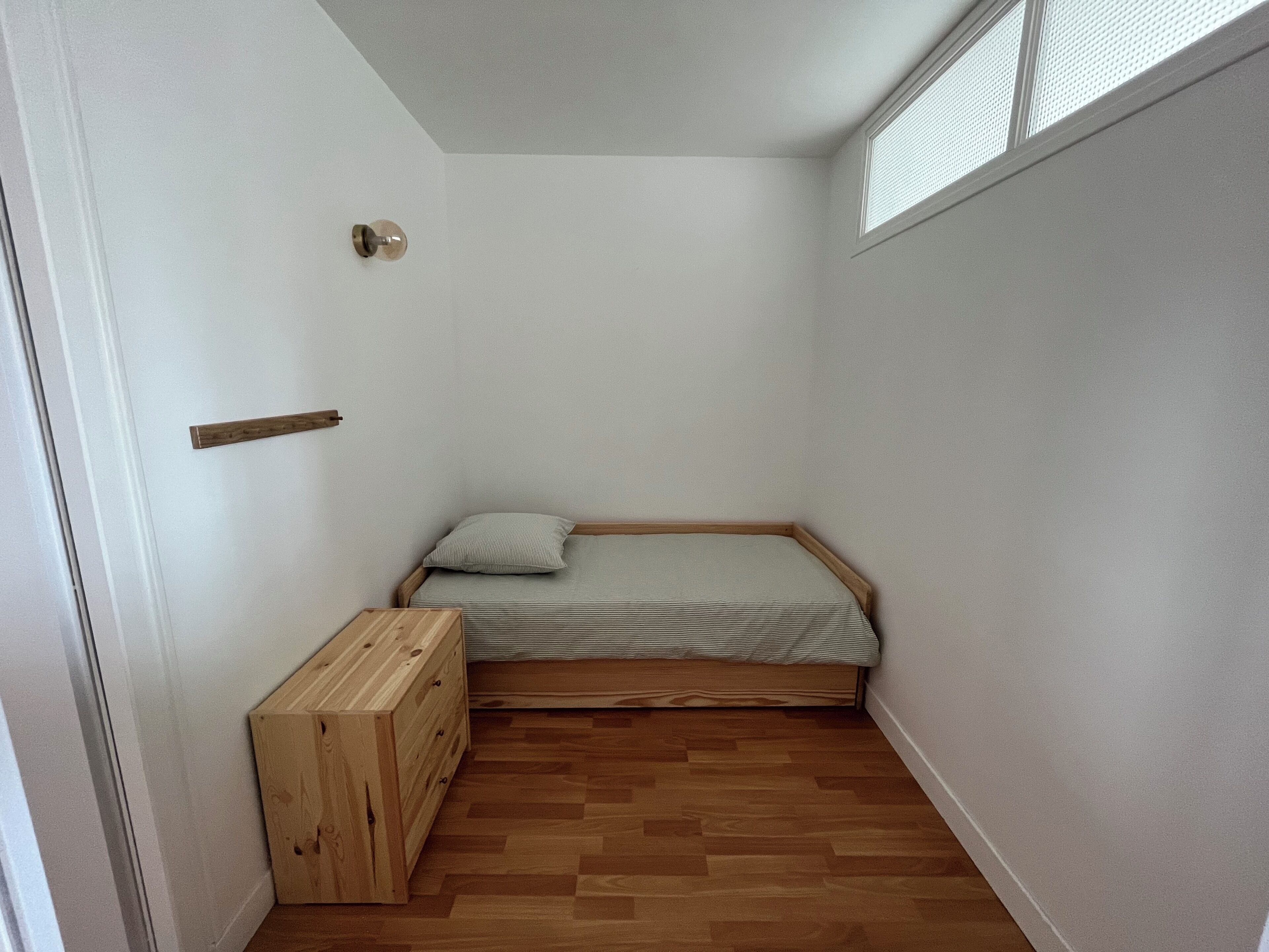 A small, single-bed room with a wooden dresser, a window, and a door.