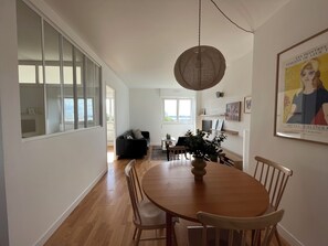 A dining area with a round wooden table, chairs, a hanging light fixture, and a view through a glass door to a balcony. - Le Congrès - Cozy apartment 50 meters from the beach (Royan)