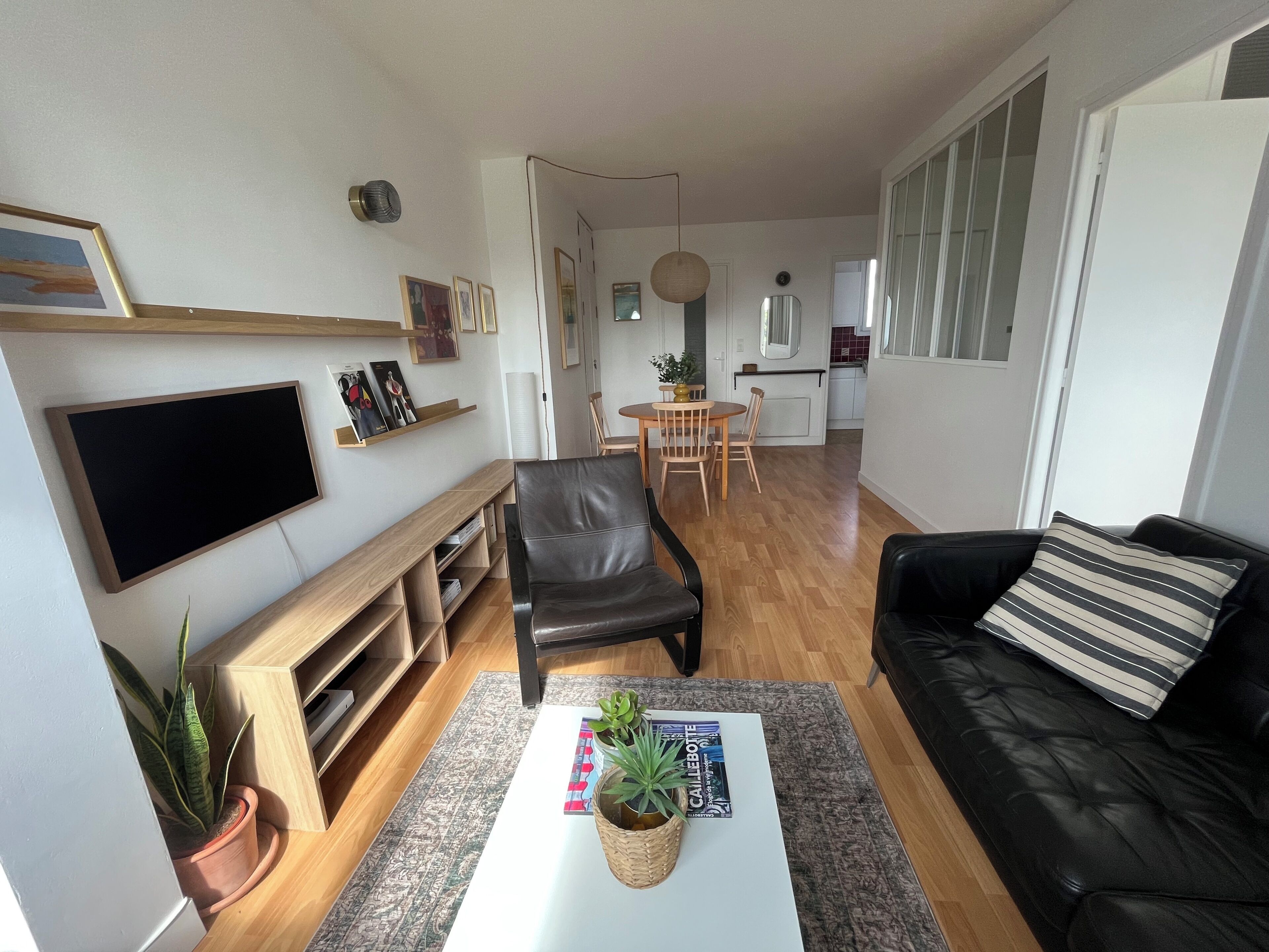 Una sala de estar moderna con un sofá de cuero negro, una mesa de centro blanca con revistas y plantas, un televisor montado en la pared y un comedor con una mesa y sillas de madera.