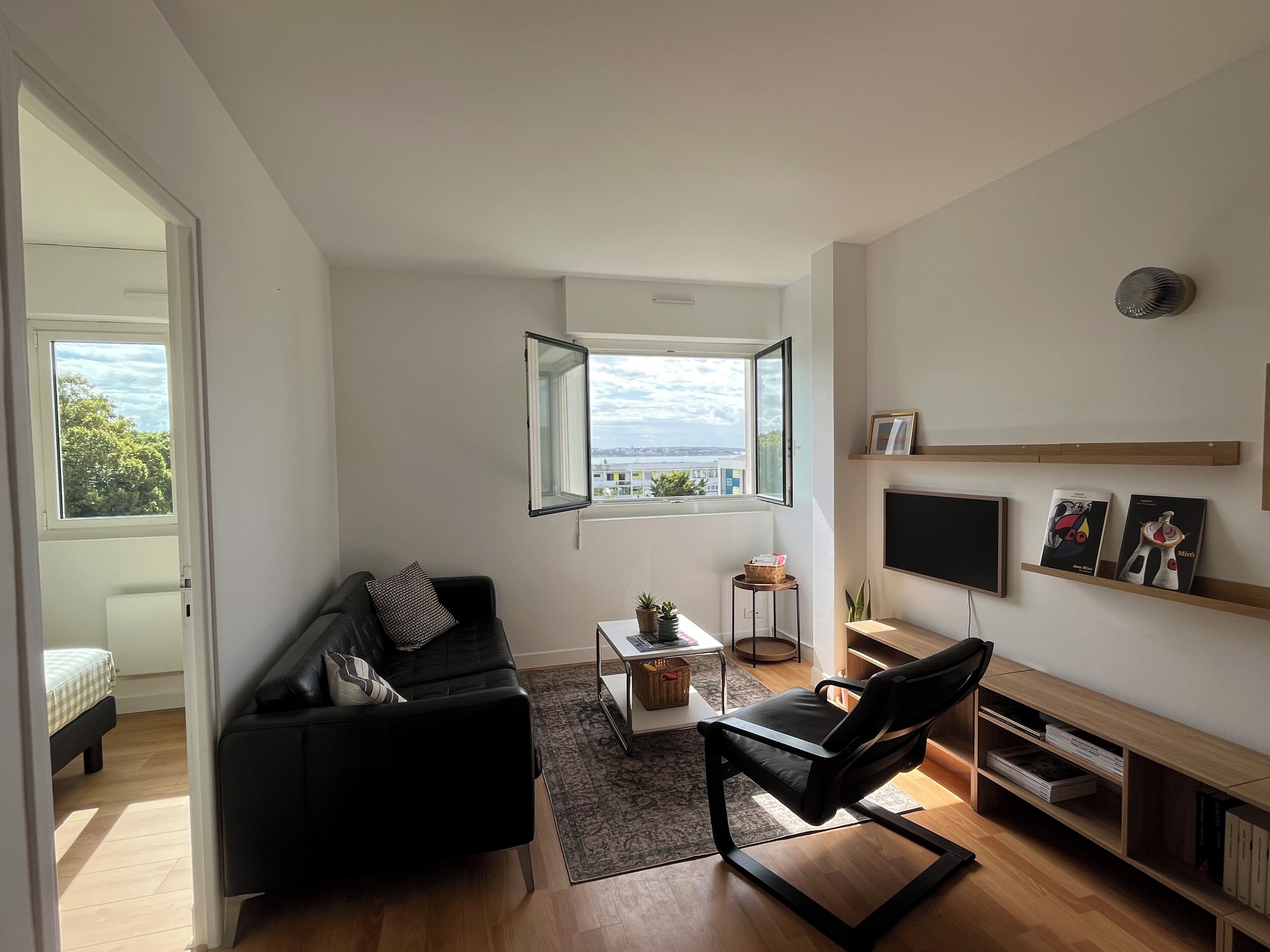 Een moderne woonkamer met een zwarte leren bank, een zwarte fauteuil, een klein bijzettafeltje, een televisie en een boekenkast.