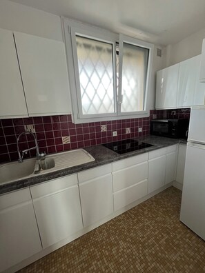 A kitchen with white cabinets, a sink, a microwave, and a window with frosted glass. - Le Congrès - Cozy apartment 50 meters from the beach (Royan)