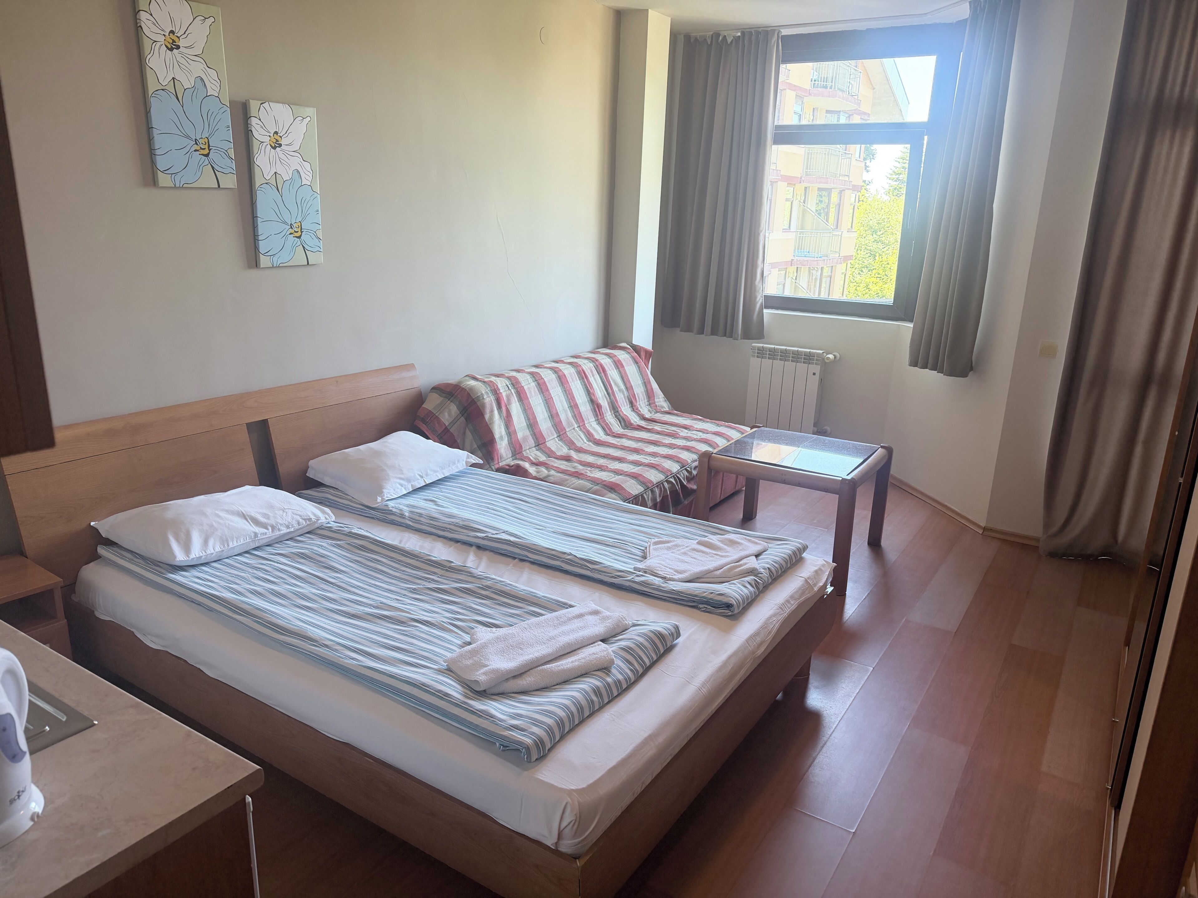 A hotel room with a double bed, a bedside table, a window with curtains, and a radiator.