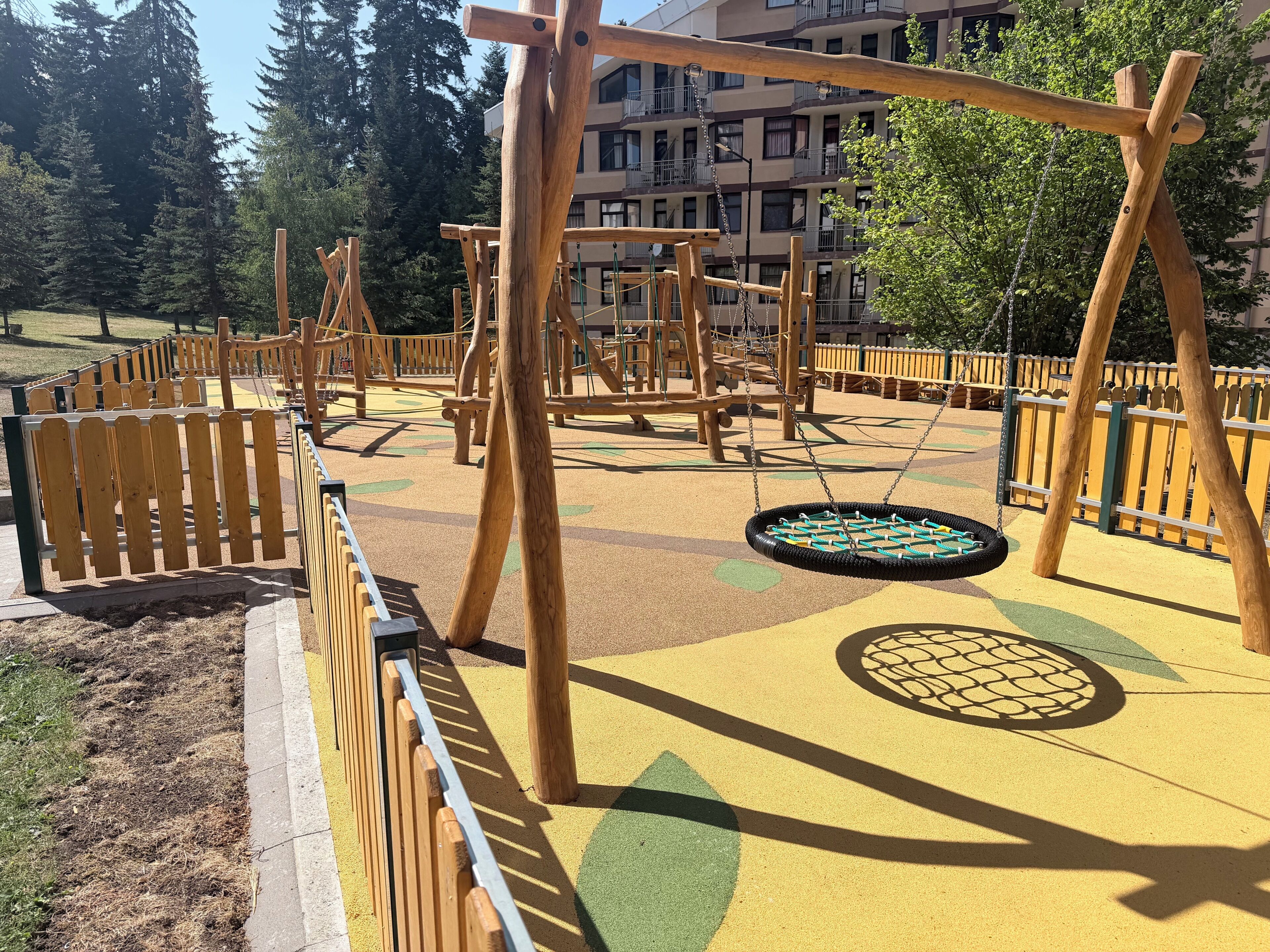 Un parque infantil con estructuras de madera y suelo de juego amarillo.