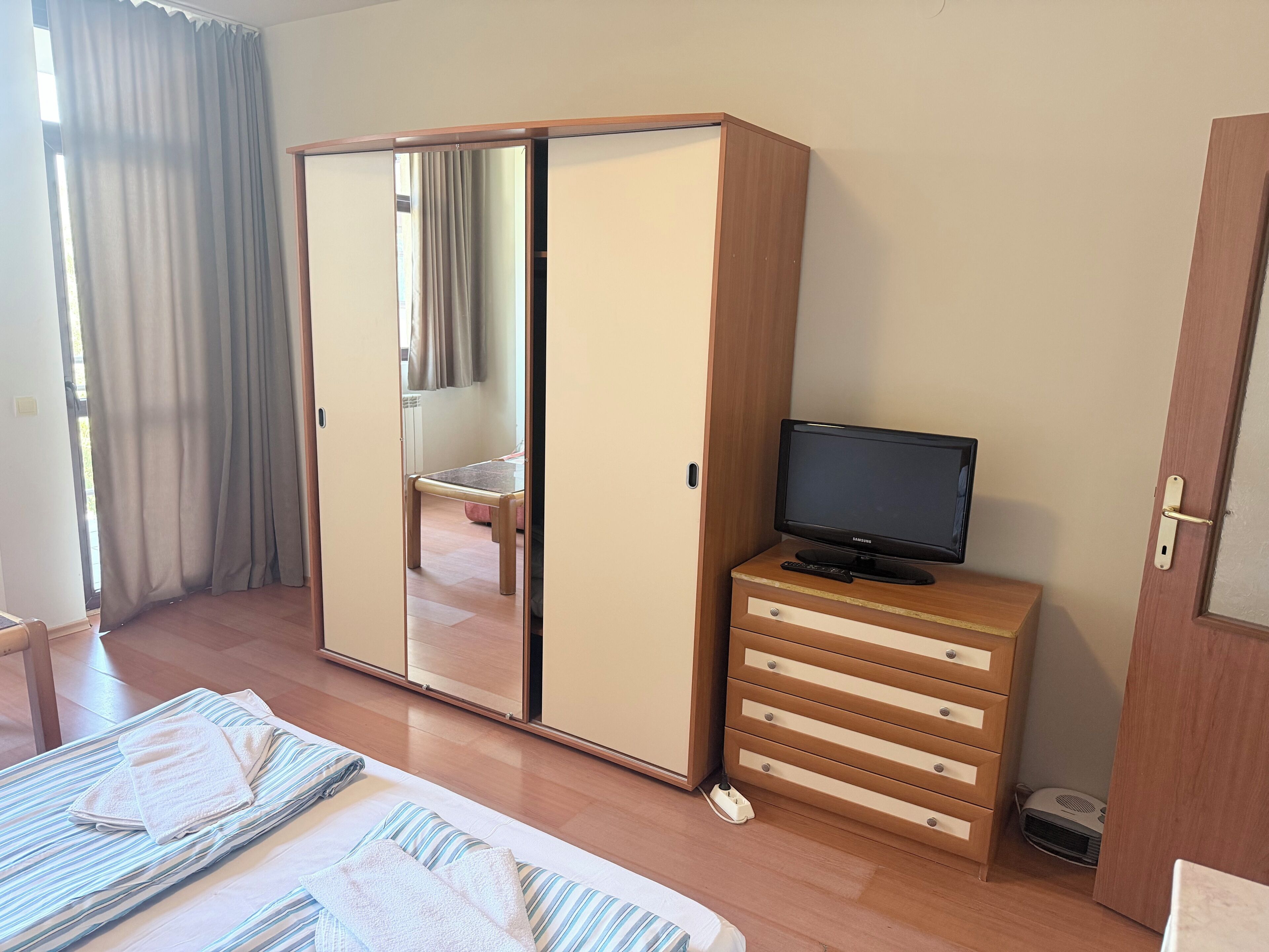 Un dormitorio con un armario, un televisor sobre una cómoda, una cama con toallas y una ventana con cortinas.
