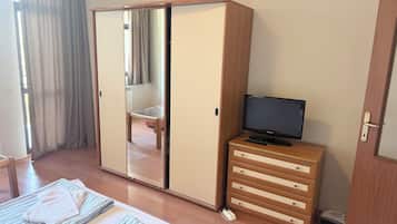 Un dormitorio con un armario, un televisor sobre una cómoda, una cama con toallas y una ventana con cortinas.