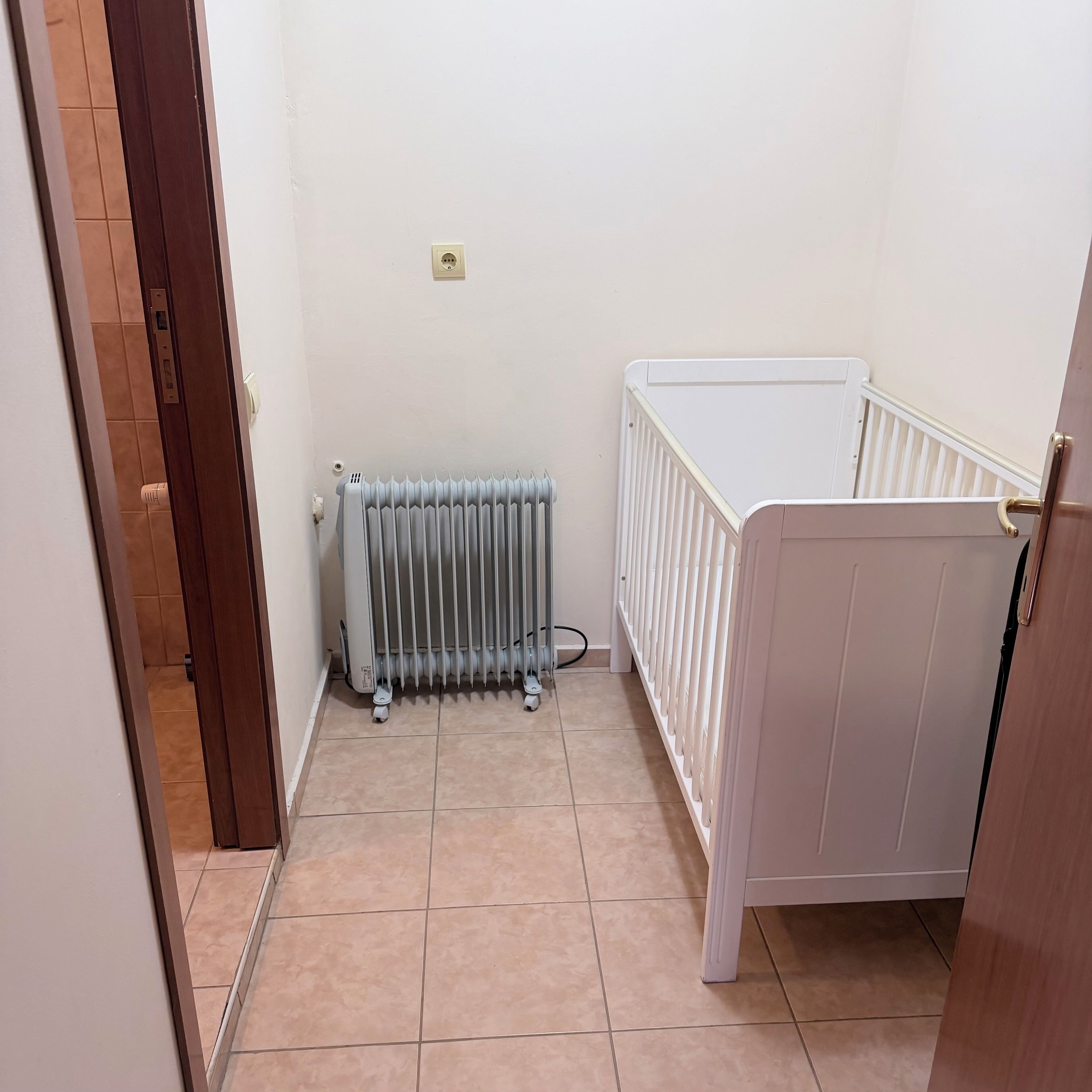 Quarto com um radiador branco, um berço branco e uma porta que dá para outro cômodo.