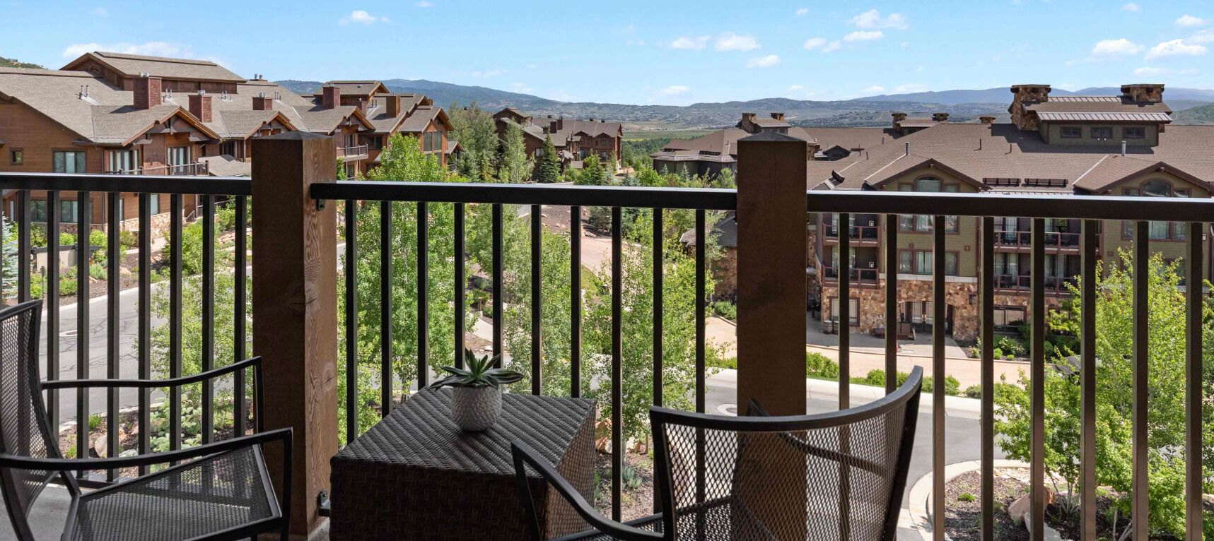A balcony with a table and chairs, overlooking a residential area with houses and a mountain range in the background.