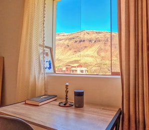 A room with a window overlooking a mountainous landscape, a framed picture on the wall, a book, a candle, and a small statue on a table.