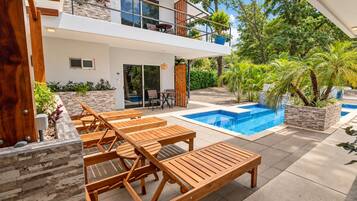 Una casa moderna de dos pisos con piscina, terraza de madera y mobiliario exterior.