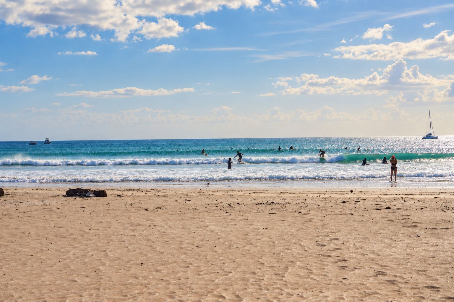 Una playa con gente practicando surf y nadando, barcos en el agua y un velero a lo lejos.