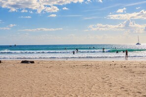 Una spiaggia con persone che fanno surf e nuotano, barche nell'acqua e una barca a vela all'orizzonte.