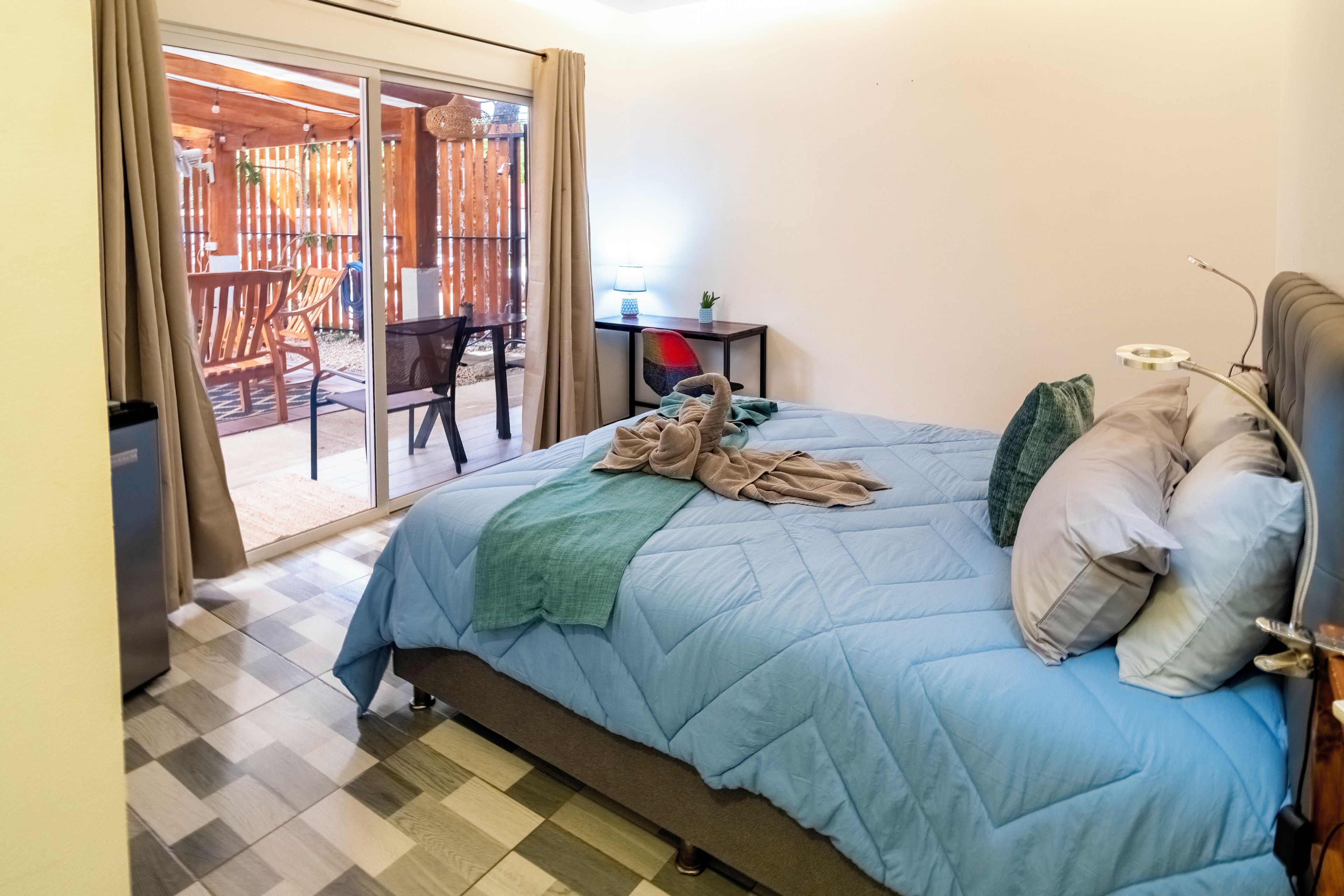 A bedroom with a bed, a chair, a table, and a view of an outdoor area with a wooden deck and chairs.