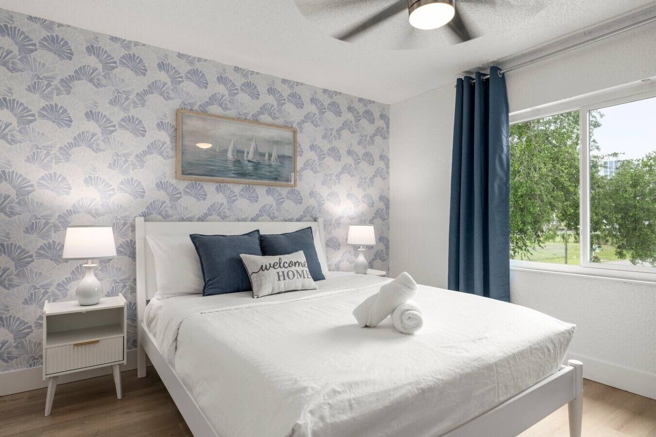 A bedroom with a bed, two bedside tables, a ceiling fan, a window with curtains, and a wall with a floral pattern.