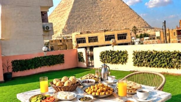 Mesa de jantar ao ar livre com comida e bebidas, uma pirâmide ao fundo e um edifício com varanda.