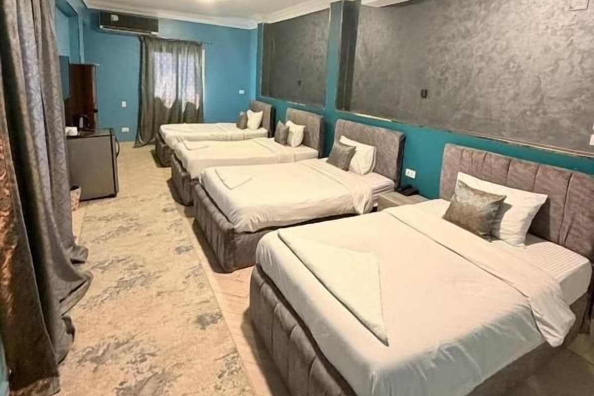 Ein Hotelzimmer mit vier Betten, die nebeneinander stehen, einem Schreibtisch und einem großen Fenster mit Vorhängen.