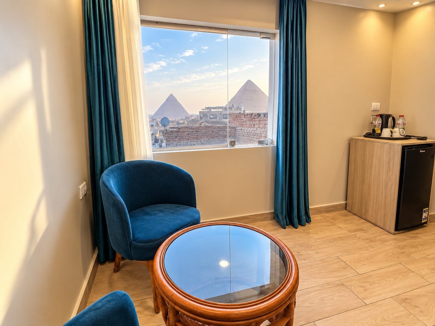 Habitación de hotel con vista a las pirámides, un sillón azul, una mesita de vidrio y un minibar.